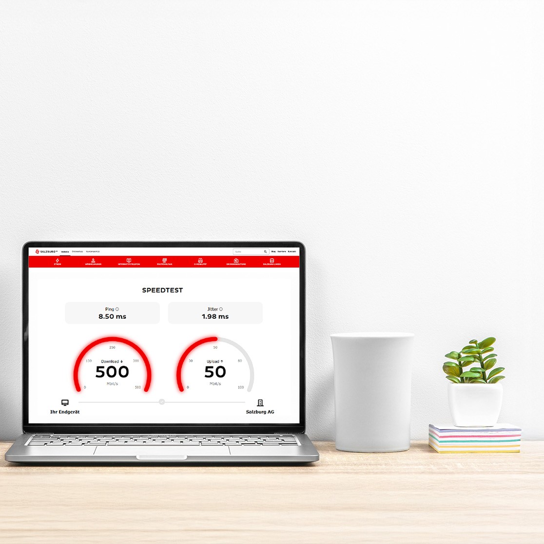 Ein Laptop steht auf einem Holzschreibtisch vor einer weißen Wand. Auf dem Bildschirm ist ein Internet-Geschwindigkeitstest mit rotem Interface zu sehen. eben dem Laptop befinden sich zwei kleine Topfpflanzen, eine weiße Tasse und ein Stapel bunter Notizzettel.