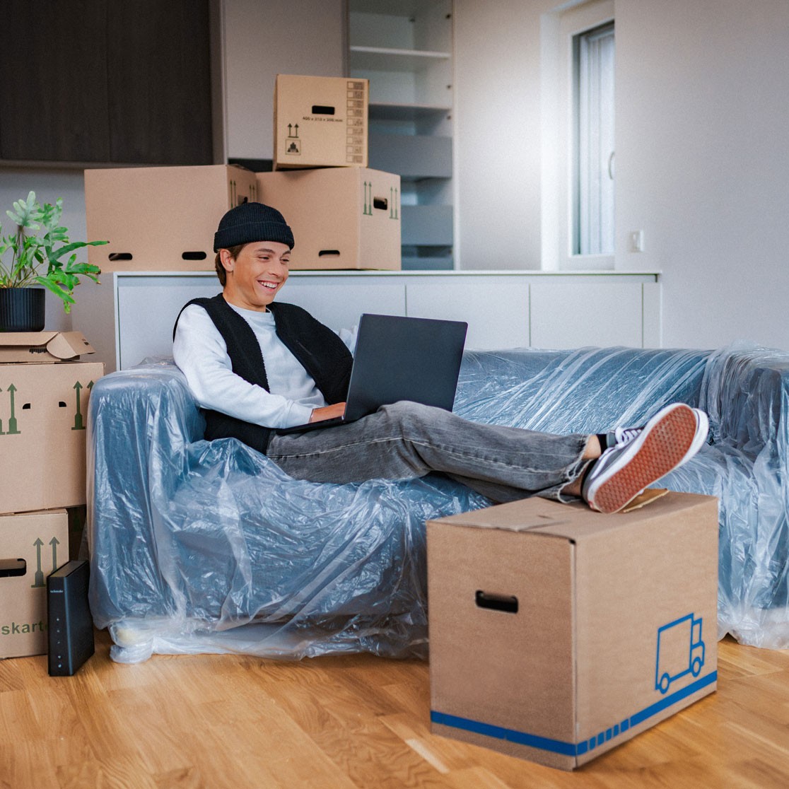 Junger Mann sitzt zwischen Umzugskartons auf einer noch abgedeckten Couch und surft am Laptop.