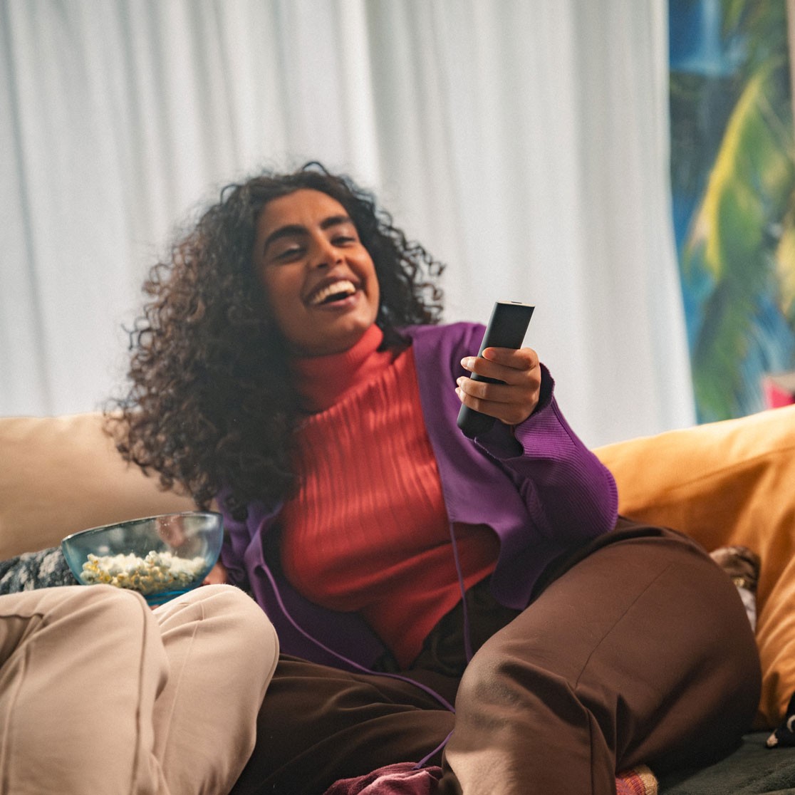 Lachende Frau auf Sofa mit Fernbedienung bei einem Fernseherlebnis.