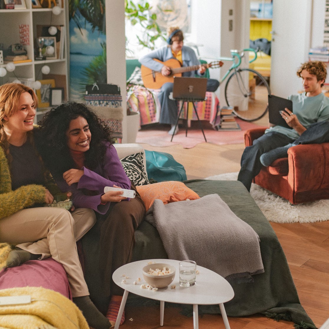 Eine bunte WG mit 4 Personen. Zwei sitzen auf einem Sofa mit Fernbedienung, schräg dahinter sitzt ein Mann mit Tablet, ihm Hintergrund spielt eine Frau Gitarre vor dem Laptop.