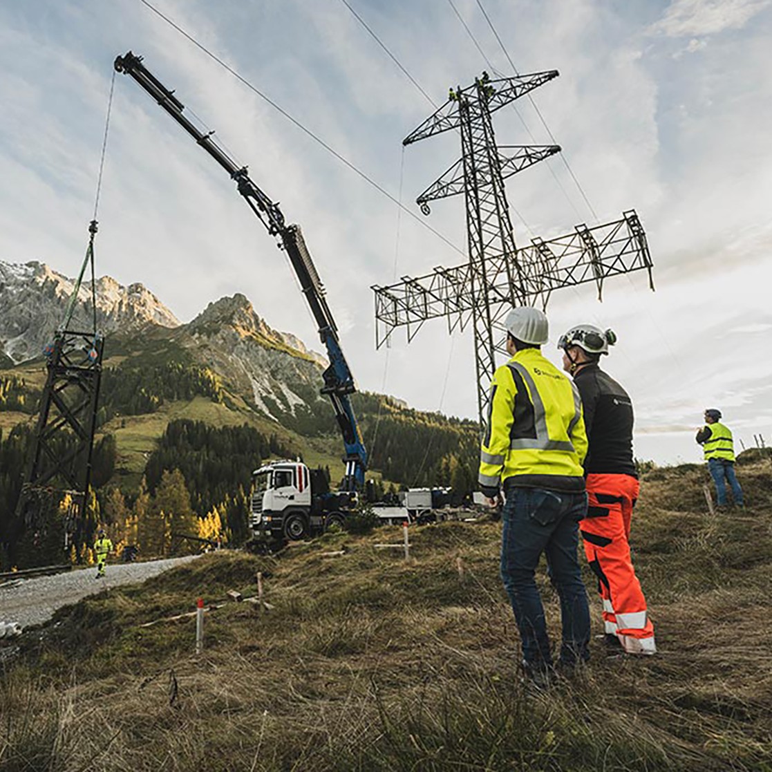 Hochspannungsmast-Montage mit Kran in Berglandschaft.