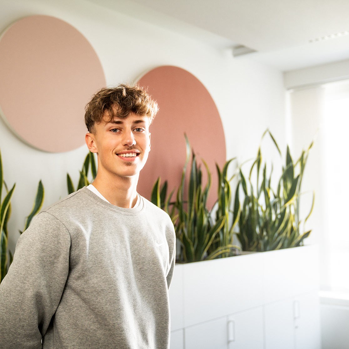 Junger Mann steht in modernen Büro mit Pflanzen im Hintergrund und blickt lächelnd in die Kamera