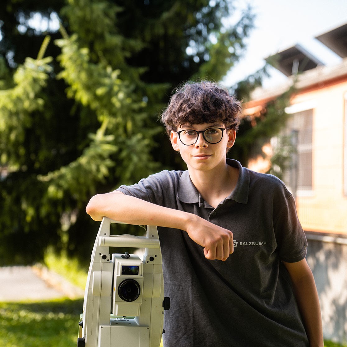 Lehrling der Salzburg AG lehnt lässig auf einem Vermessungsgerät mit Tannen und Gebäude im Hintergrund