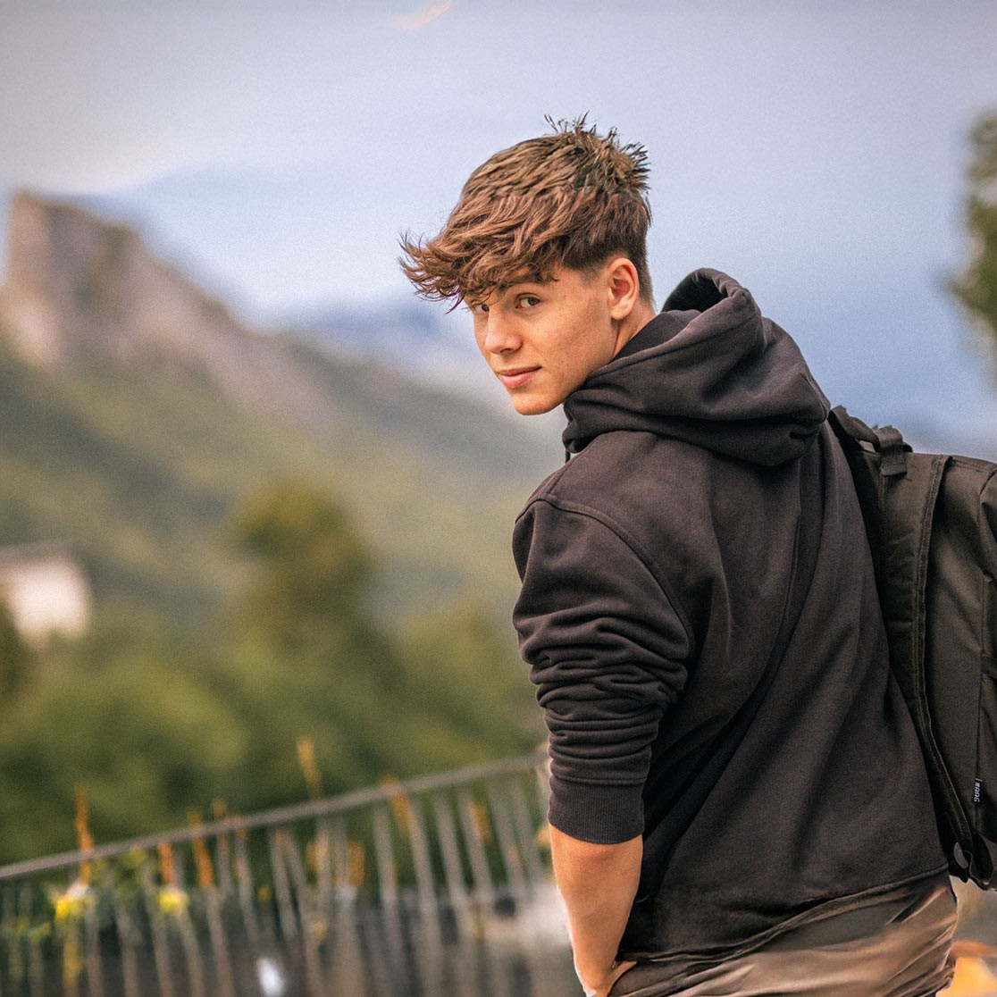EIn Junge im Schwarzen Pullover steht vor einer Berg Kulisse.