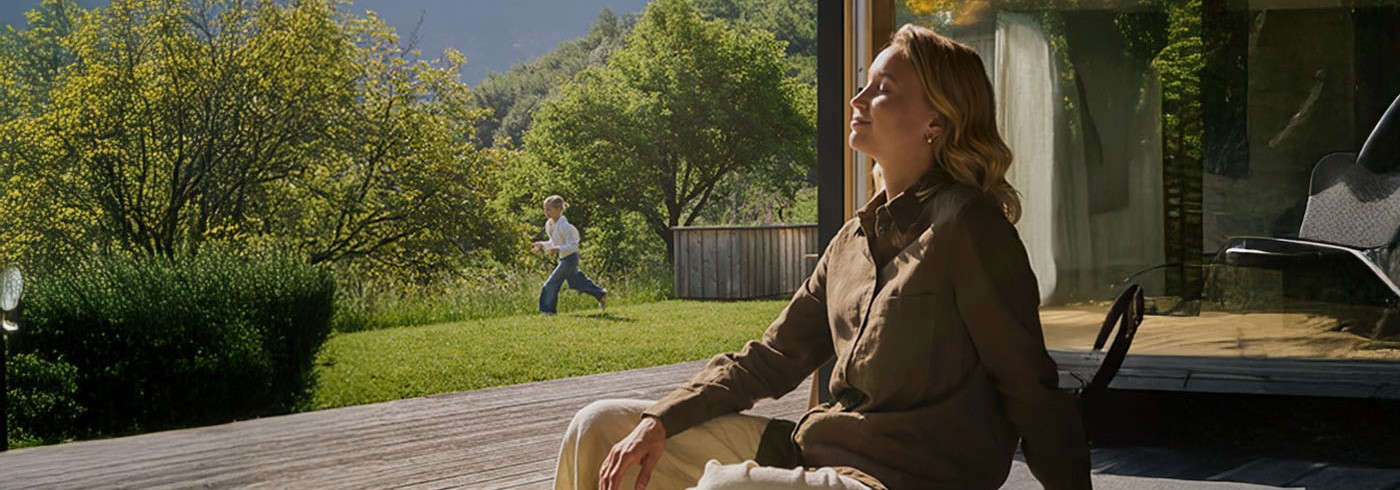 Eine Frau sitzt auf der Terrasse und genießt die Sonne.  Im Hintergrund läuft ein Kind durch den Garten.