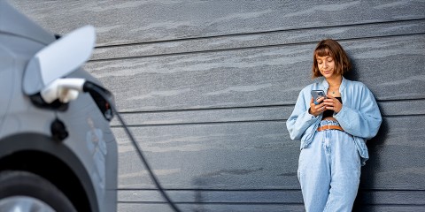 Frau lehnt neben ladendem E-Auto an der Wand und bedient Smartphone.