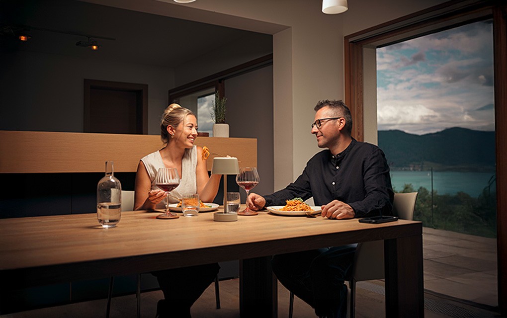 Zwei Personen sitzen an einem Holztisch in einem modernen Essbereich und essen Pasta. Auf dem Tisch stehen zwei Gläser Rotwein, eine Wasserflasche und eine kleine Lampe. Im Hintergrund ist durch eine große Glasfront ein Blick auf einen See und umliegende Berge zu sehen.