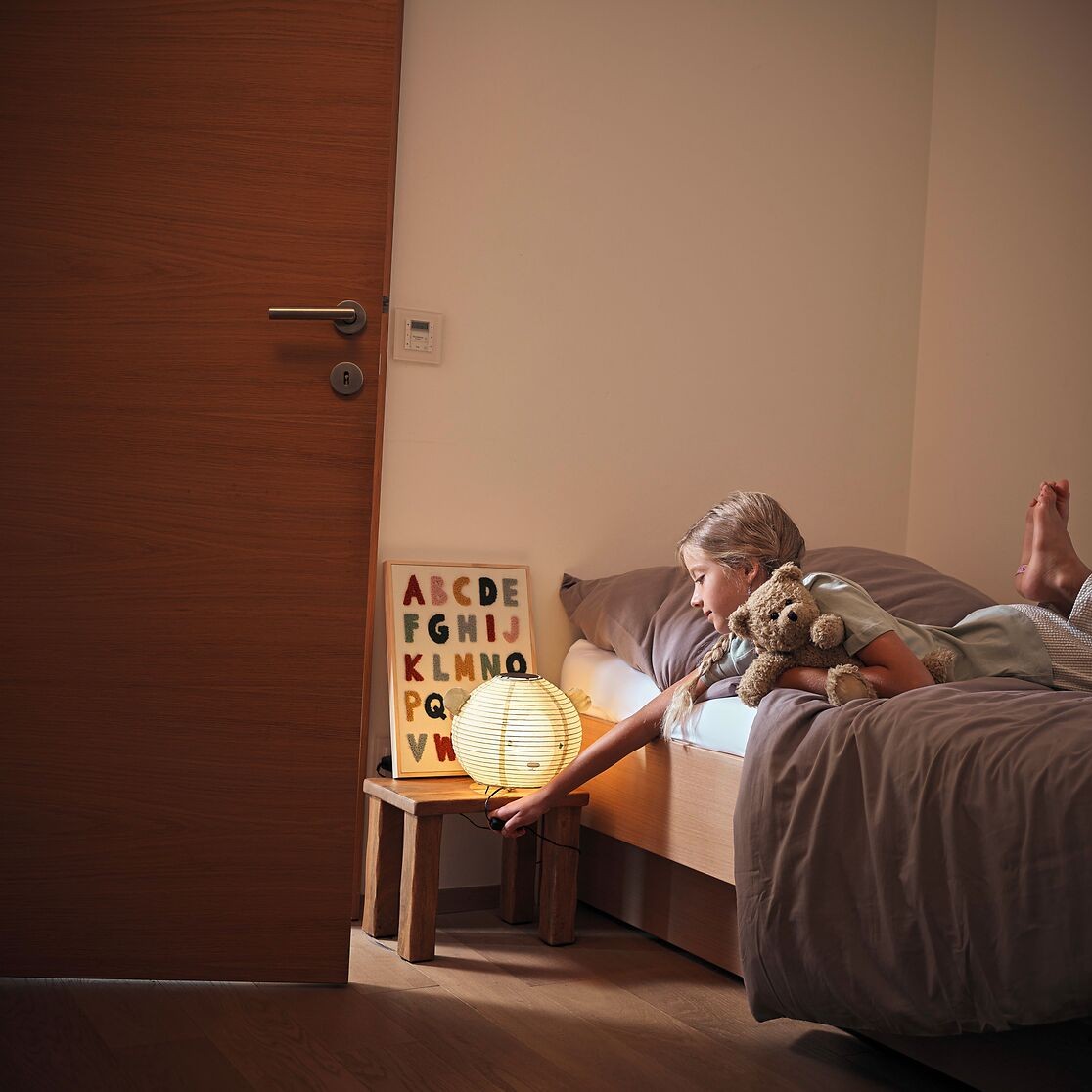 Mädchen liegt auf Bett mit Teddybär und knipst Licht aus.